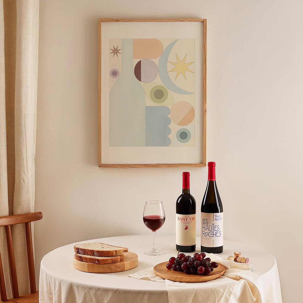 Two bottles of wine on a table with a glass, grapes, and bread, with a framed abstract art piece on the wall.